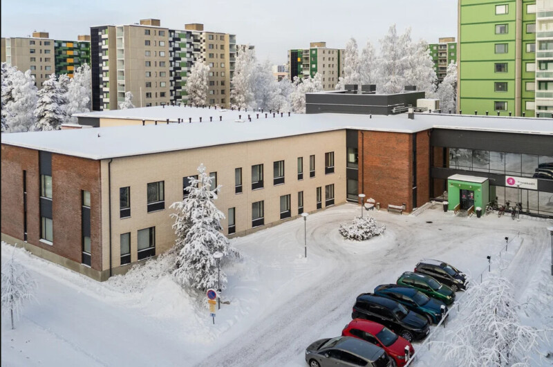 Kaksikerroksinen rakennus, edessä parkkipaikka ja takana kerrostaloja, ympärillä luminen maisema.