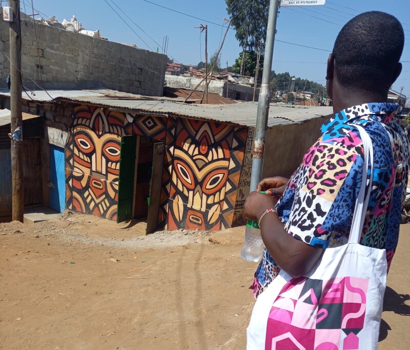 Lyhythiuksinen ihminen seisoo matalan, värikkään rakennuksen edessä selkä päin kameraa, olalla Rinnekotien kangaskassi.