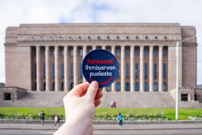 Henkilö pitää kädessään Rohkeasti ihmisarvon puolesta -merkkiä, taustalla Suomen eduskuntatalo.