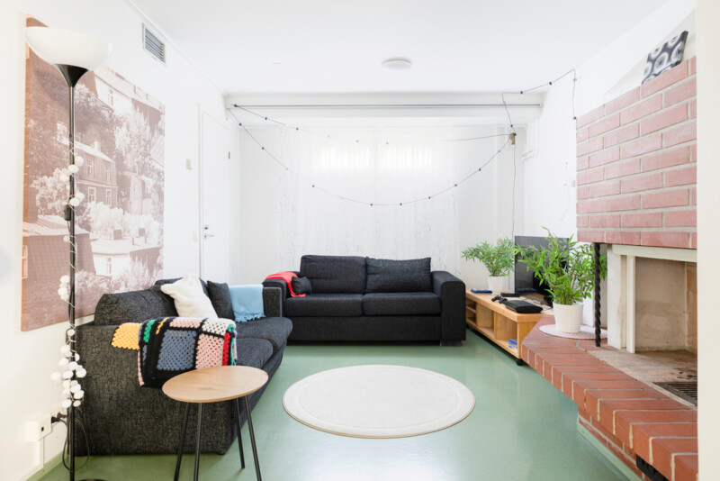 Oleskelutilassa yhdellä seinustalla avonainen takka ja pieni tv-taso, jonka päällä viherkasveja ja tv. Toisella seinustalla kaksi mustaa sohvaa.