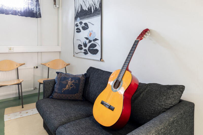 Tumman sohvan päällä klassinen kitara. Nurkassa kaksi puista tuolia, seinällä seinäryijy.