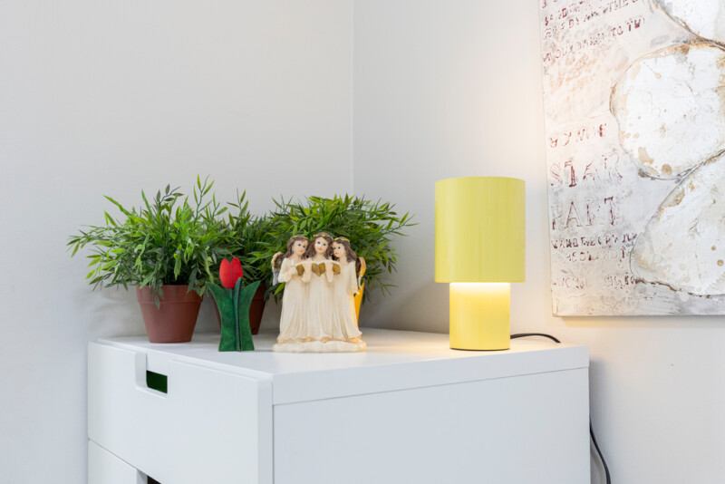 Valkoisen lipaston päällä viherkasveja, keltainen pöytälamppu sekä koriste-esineitä. Seinällä vaaleanhohtoinen taulu.