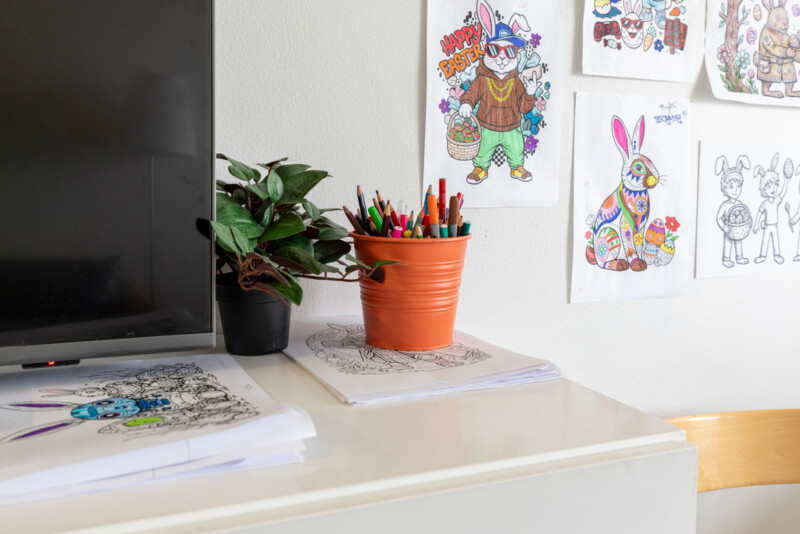 Hyllyn päällä pinoissa piirroskuvia, niiden päällä astiassa värikyniä. Seinälle kiinnitetty puputeemaisia värityskuvia.