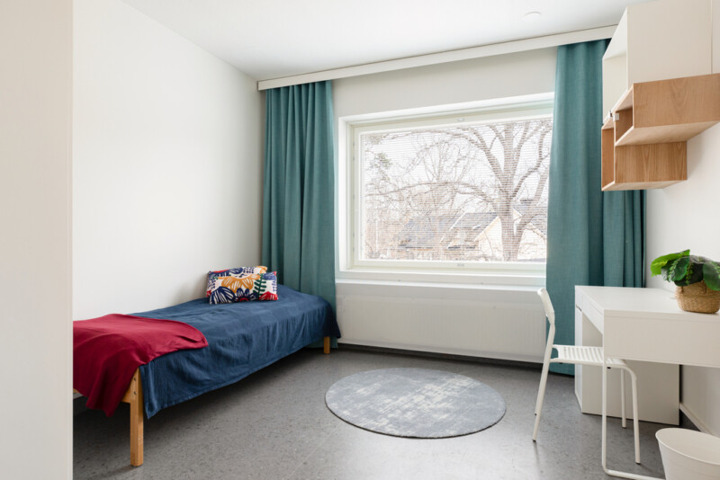 Kuva huoneesta, jonka yhdellä seinällä pedattu sänky, toisella seinällä valkoinen kirjoituspöytä ja tuoli. Ikkunoita kehystää petrolin väriset verhot.