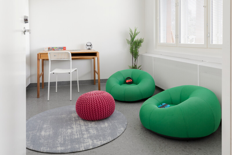 Huoneessa kaksi vihreää pehmotuolia lattialla, maton päällä myös punainen rahi. Seinän vieressä puinen kirjoituspöytä ja tuoli. Nurkassa viherkasvi.