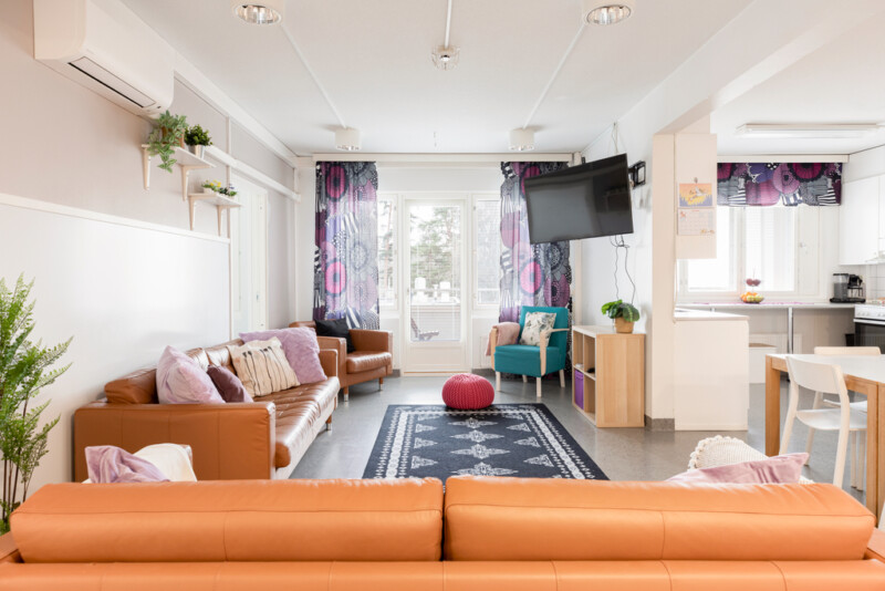Oleskelutilassa vaaleanruskeita sohvia, oikealla sivulla näkyy myös keittiötä. Oleskelutila on kodinomaisen näköinen, ikkunassa värikkäät verhot.