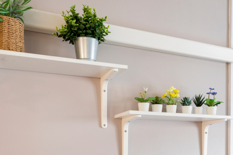 Seinällä kiinnitettyjä valkoisia hyllyjä, joissa pieniä viherkasveja ja kukkia.