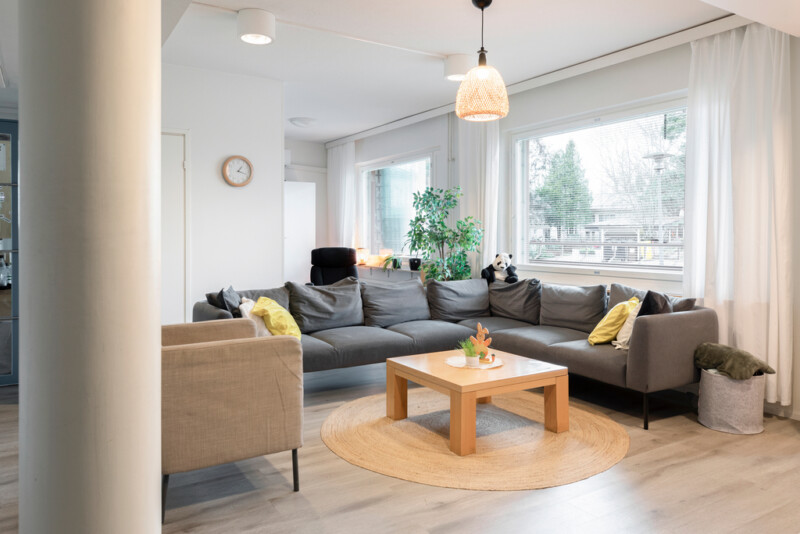 Oleskelutilassa iso tummanharmaa kulmasohva ja beige nojatuoli. Isoja ikkunoita reunustaa vaaleat läpinäkyvät verhot. Huoneessa on myös korkea viherkasvi. Huone on kodinomainen ja valoisa.