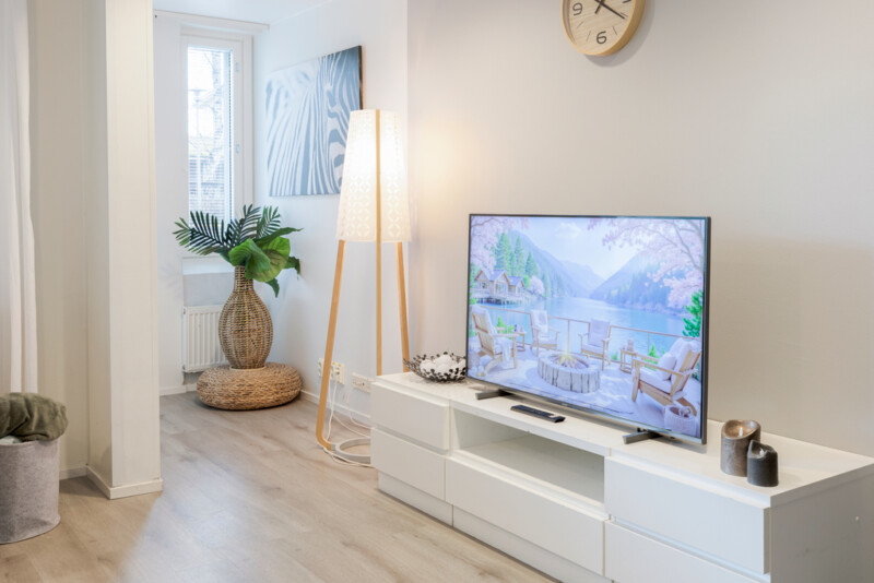 Valkoisen matalan televisiotason päällä televisio sekä kaksi kynttilää ja kulho. Tason vieressä korkeahko jalkalamppu sekä nurkassa rottinkisessa maljassa iso viherkasvi.