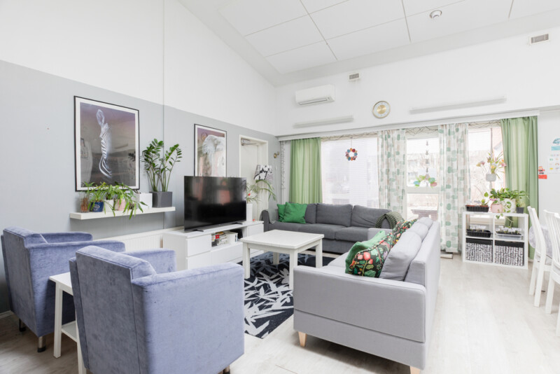 Oleskelutilassa vaaleanharmaan ja -sinisen sävyisiä sohvia ja nojatuoleja ympäröi valkoista sohvapöytää. Tilassa myös televisiotaso, jolla televisio sekä isoja tauluja seinällä. Ikkunoita kehystää vihreänsävyisiä verhoja.