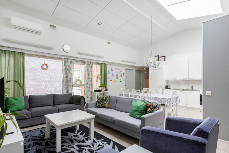 Oleskelutilassa vaaleanharmaan ja -sinisen sävyisiä sohvia ja nojatuoleja ympäröi valkoista sohvapöytää. Tilassa myös televisiotaso, jolla televisio sekä isoja tauluja seinällä. Ikkunoita kehystää vihreänsävyisiä verhoja. Taustalla näkyy keittiö, jossa iso ruokailupöytä ja sen ympärillä tuoleja.