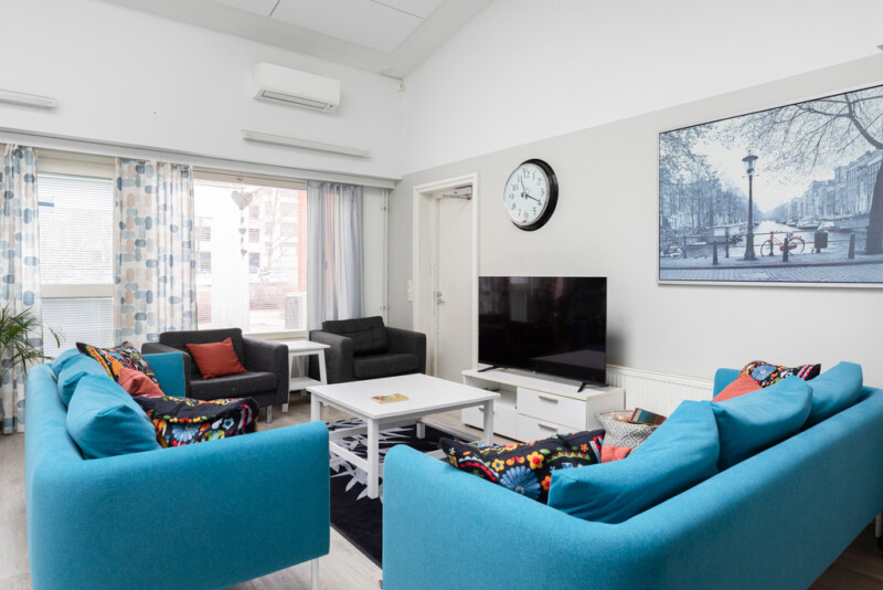 Oleskelutilassa kaksi sinistä sohvaa sekä mustia nojatuoleja. Keskellä huonetta valkoinen sohvapöytä, jonka takana televisiotaso.