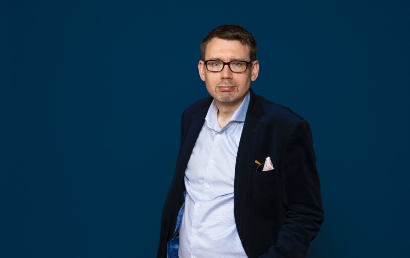 Mies puvun takki päällä katsoo kameraan, tummansininen tausta.