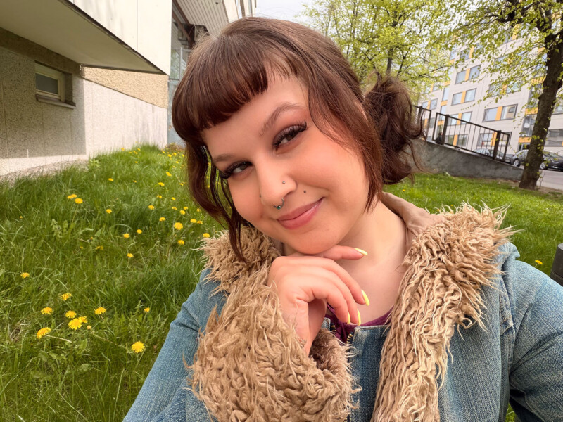 Henkilö katsoo kesäisessä kuvassa hymyillen kameraan sormet leuan alla kerrostalon takapihalla.