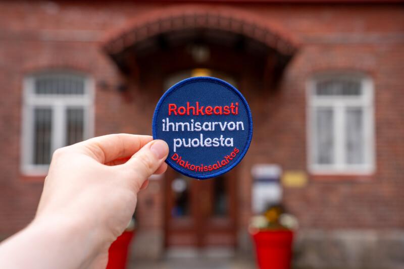 Käsi pitelee rintamerkkiä, jossa on teksti Rohkeasti ihmisarvon puolesta. Taustalla punatiilinen rakennus.