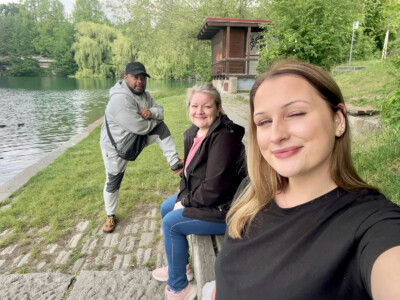 Kolme henkilöä hymyilee kesäisessä selfiekuvassa puistomaisemassa maisemassa veden äärellä.