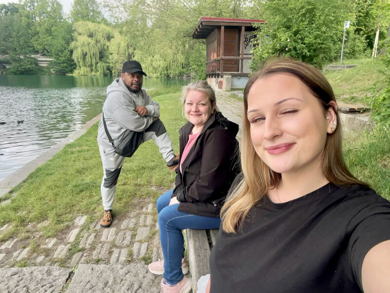 Kolme henkilöä hymyilee kesäisessä selfiekuvassa puistomaisemassa maisemassa veden äärellä. 