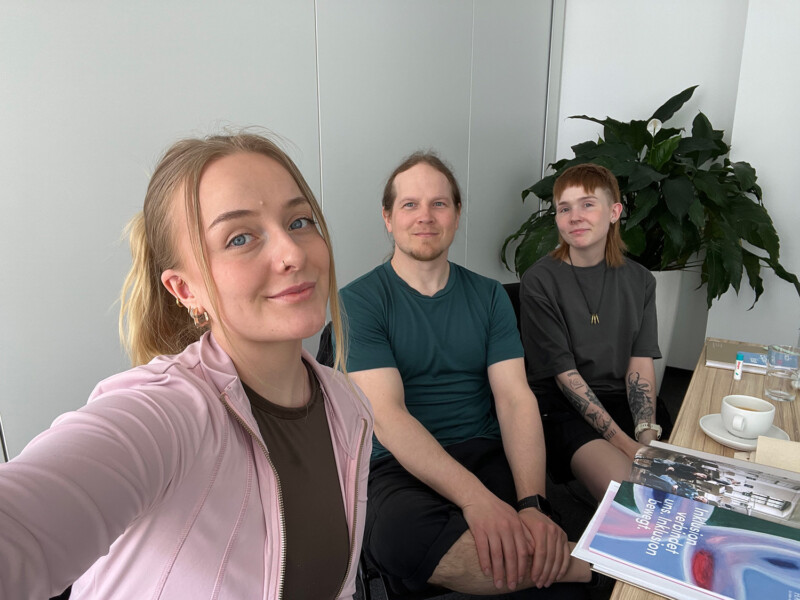 Kolme hymyilevää henkilöä istuu selfiekuvassa pöydän äärellä, pöydällä on kahvikuppi ja printtimateriaalia.