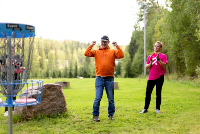 Kaksi miestä frisbeegolfkentällä, etummainen tuulettaa, takimmaisella on Rinnekotien pinkki työpaita päällä.