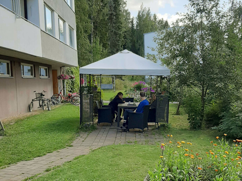 Ihmisiä kerrostalon takapihalla puutarhakatoksen alla istumassa pöydän äärellä.