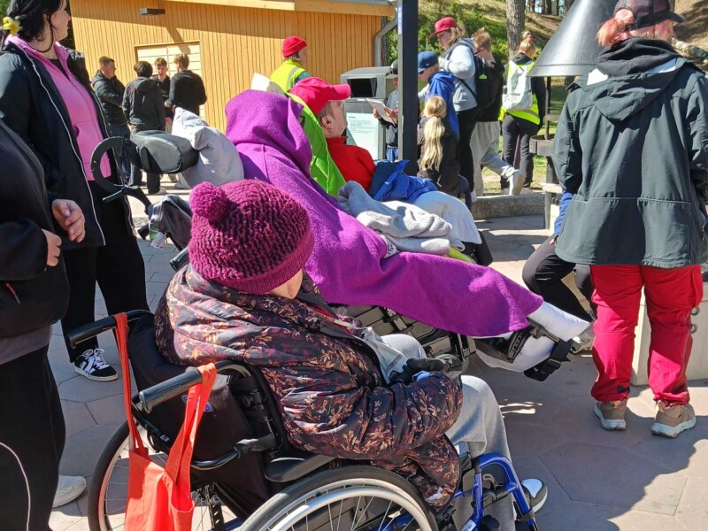 Ihmisiä pyörätuolissa ja kävellen ulkona katselemassa jotakin tapahtumaa.