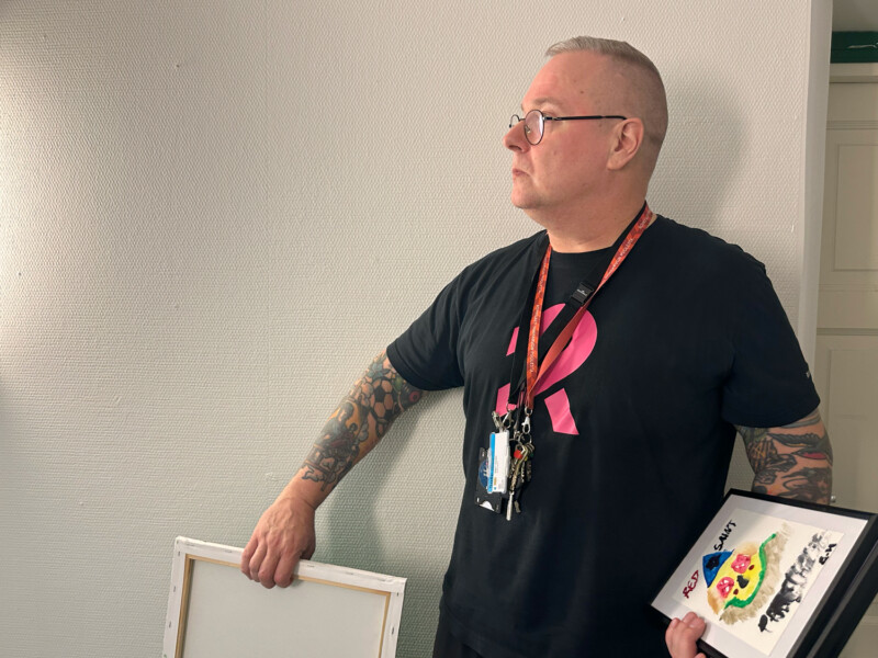Mustapaitainen silmälasipäinen mies pitää kädessään tauluja ja katsoo vasemmalle.