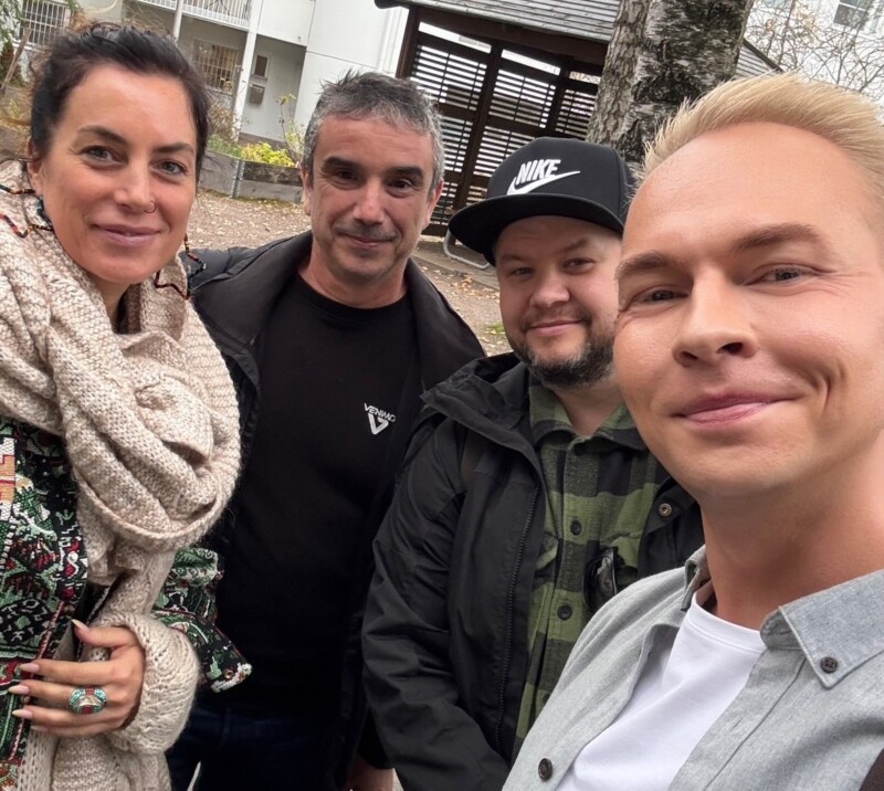 Neljä henkilöä hymyilee selfiekuvassa pihalla kerrostalon edessä.