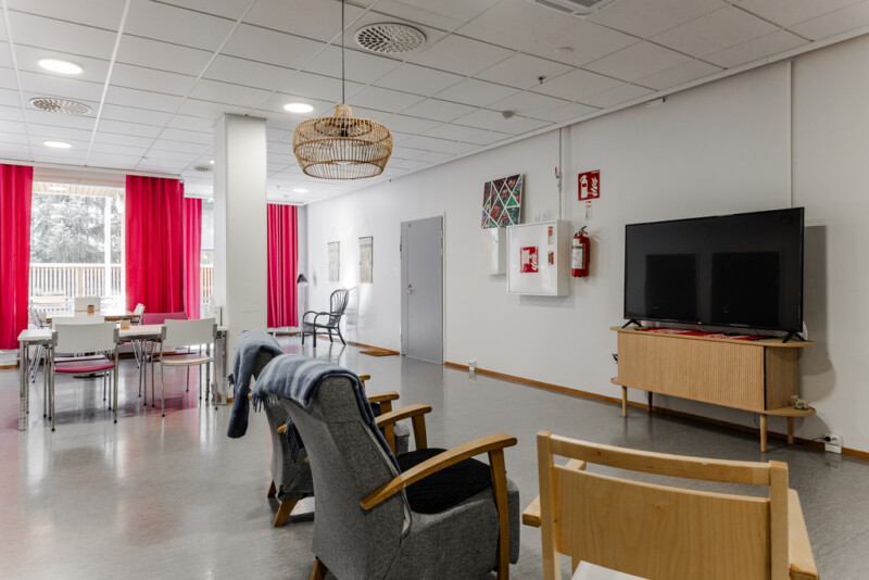 Yhteinen oleskelutila, jossa on nojatuoleja, televisio ja suuret ikkunat punaisine verhoineen.
